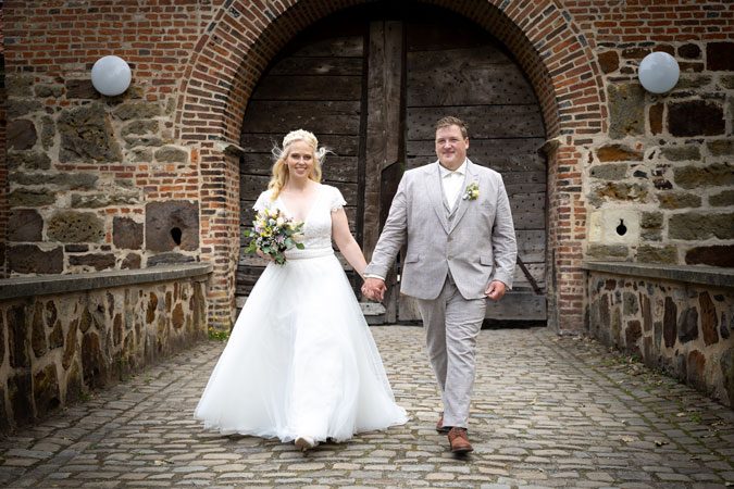 Ein Foto, dass ein Hochzeitspaar vor dem Tor, des Schoss Sythen, in Haltern am See, zeigt. Der Bräutigam trägt einen grauen Anzug und die Braut ein traditionelles weißes Kleid. Das Foto wurde von dem Fotografen Manuel Overwien aufgenommen.